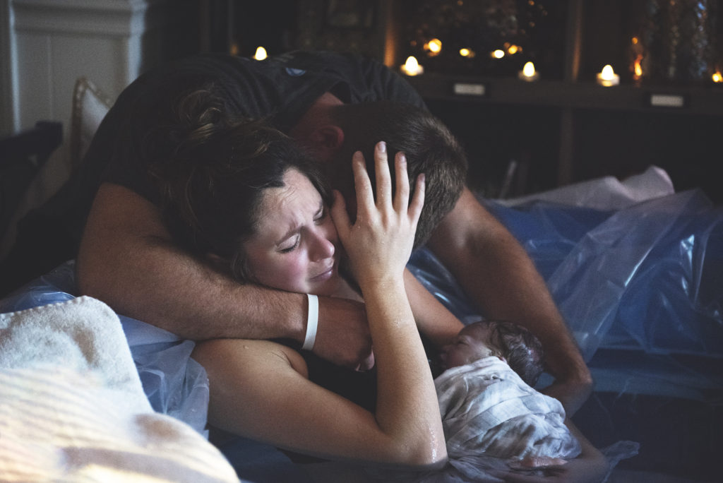 Couple embracing holding baby that was born second prior. Woman is sitting in birthing tub while husband is holder her from outside the tub. Rates page place holder.