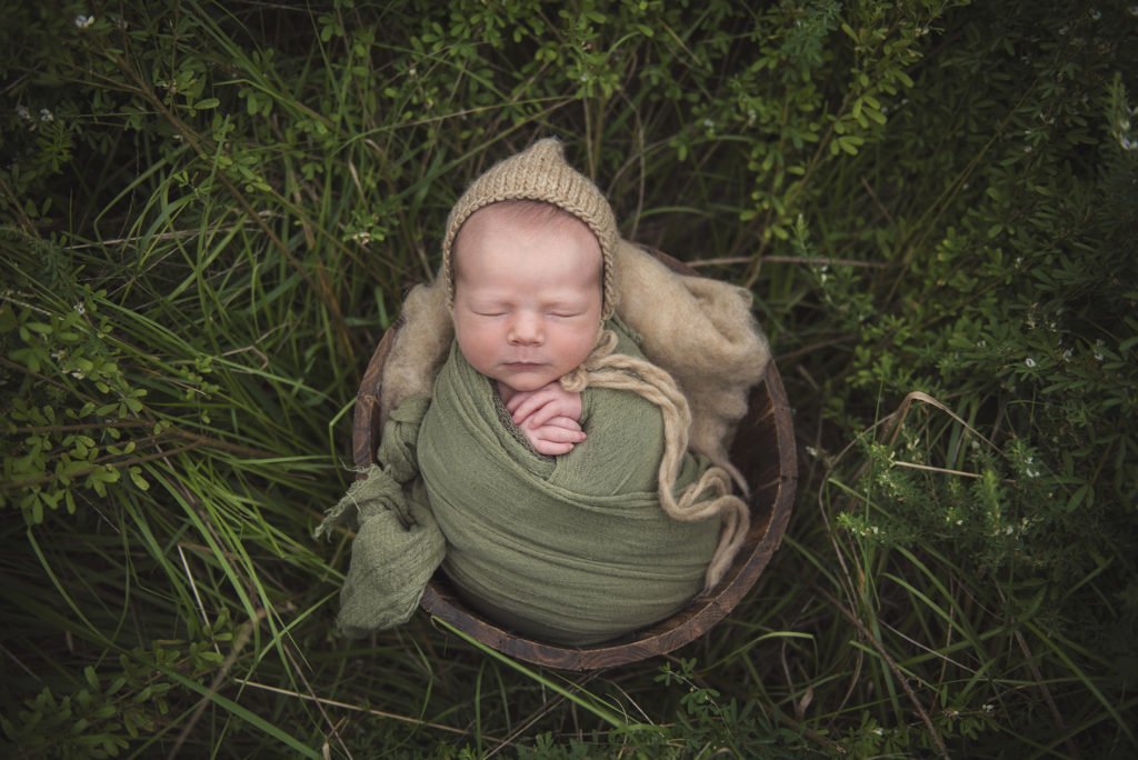 Newborn boy wrapped up sleeping with bonnet lying propped up in a bucket in a field of tall grass. Frequently Asked Questions page place holder.