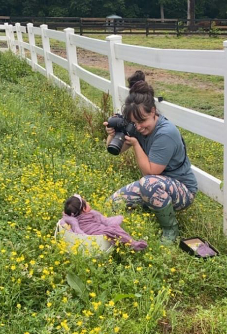Female photographer outside in a kneeling position taking a photograph of a newborn girl in a prop located in a field of buttercups.