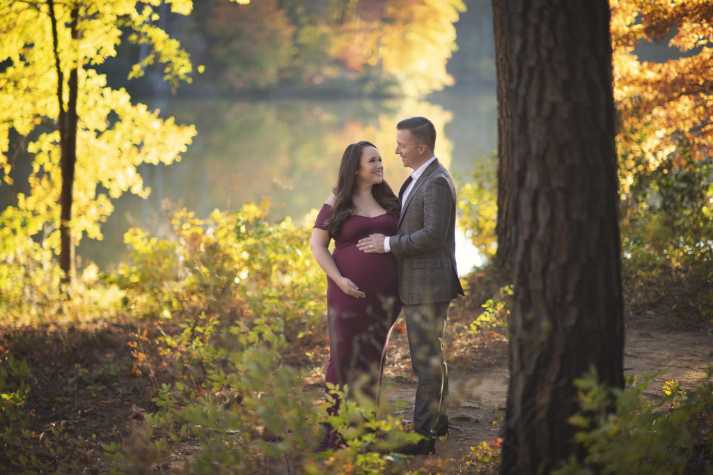 Pregnant woman and husband standing in the woods in front of a lake looking at each other.